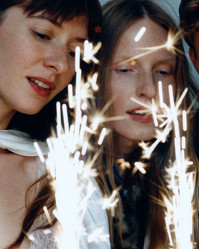 Two adult females in an indoor environment, looking at bright sparklers. The women have light skin and are wearing casual clothing.