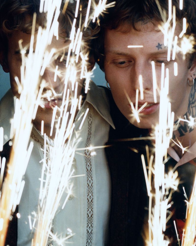 Close-up of two young adult men with light hair surrounded by bright sparks, wearing formal shirts and dark jackets in a dimly lit space.