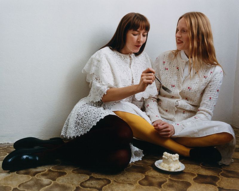 Two young women share a cake while seated on the floor. One holds a fork with cake, both wear white dresses in an indoor setting.