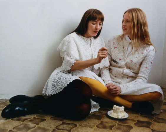 Two young women share a cake while seated on the floor. One holds a fork with cake, both wear white dresses in an indoor setting.
