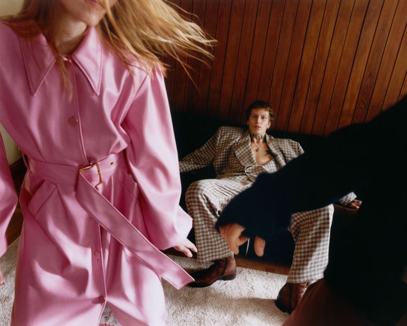Fashion shot of a blonde model wearing a pink trench coat, another model in a plaid suit, and a dark figure in an indoor setting.
