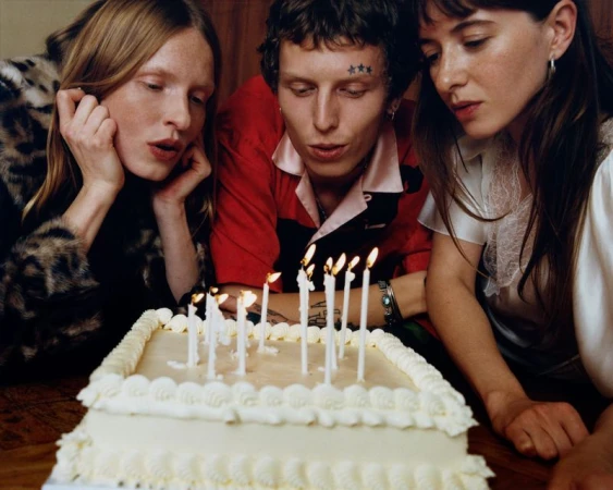 Three young adults gathered around a two-tiered cake with lit candles, blowing out the flames, in an indoor setting, medium shot.