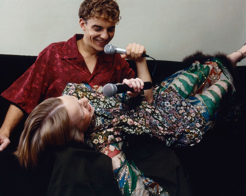 Two singers perform with microphones. One is lying down with floral pants and a floral top, while the other is standing and singing.