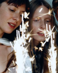 Close-up of two women looking forward as bright sparks from a handheld sparkler illuminate the foreground in a dimly lit setting