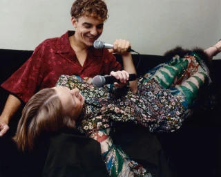 Two people holding microphones while lounging together on a dark sofa in a minimalist room
