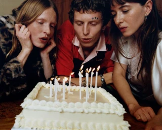 Three young adults looking down closely at a lit birthday cake with white frosting on a wooden table.