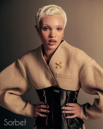 Portrait of a woman with short platinum hair wearing a beige jacket over a black corset, hands on hips, against a neutral background.