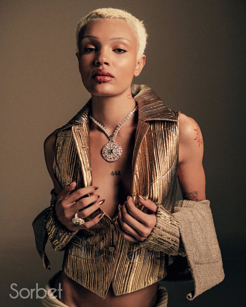 Close-up studio shot of a female model with platinum blonde hair, wearing a gold vest and jewelry, posing against a neutral background.