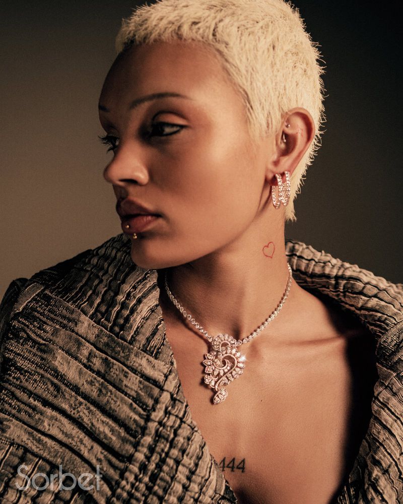 Close-up studio shot of an adult female model with short blonde hair wearing a patterned coat and diamond earrings and necklace.