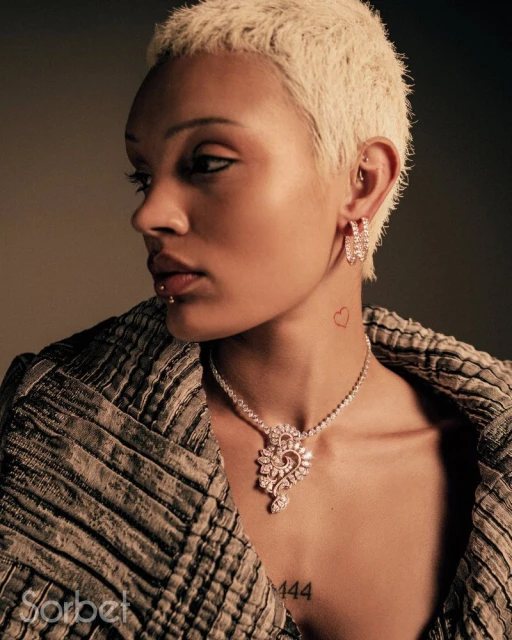 Close-up studio shot of an adult female model with short blonde hair wearing a patterned coat and diamond earrings and necklace.