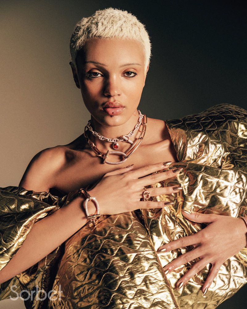 Close-up portrait of an adult female with a light complexion, platinum blonde hair, and wearing gold statement jewelry and clothing.