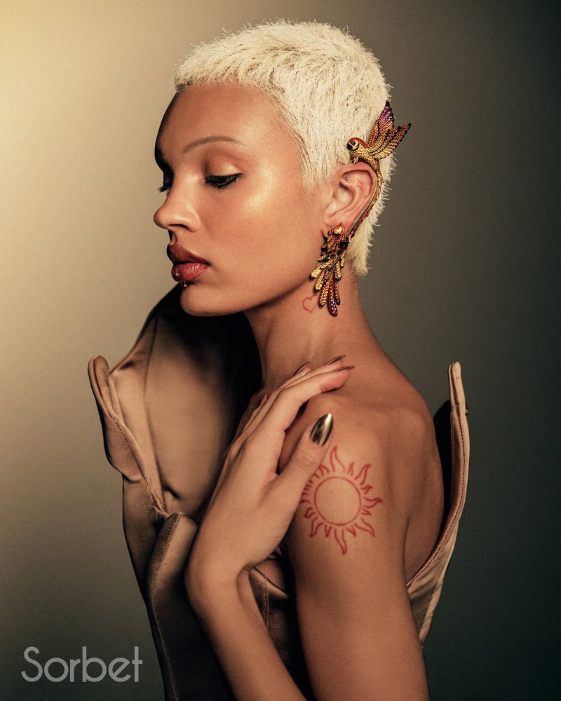 Adult woman with blonde pixie cut and bird earring in a studio setting, wearing a beige dress and hand placed on shoulder.