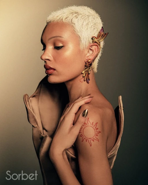 Adult woman with blonde pixie cut and bird earring in a studio setting, wearing a beige dress and hand placed on shoulder.