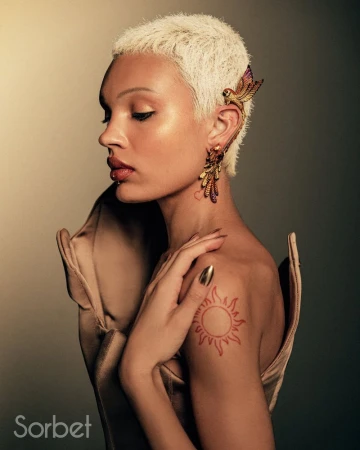 Adult woman with blonde pixie cut and bird earring in a studio setting, wearing a beige dress and hand placed on shoulder.