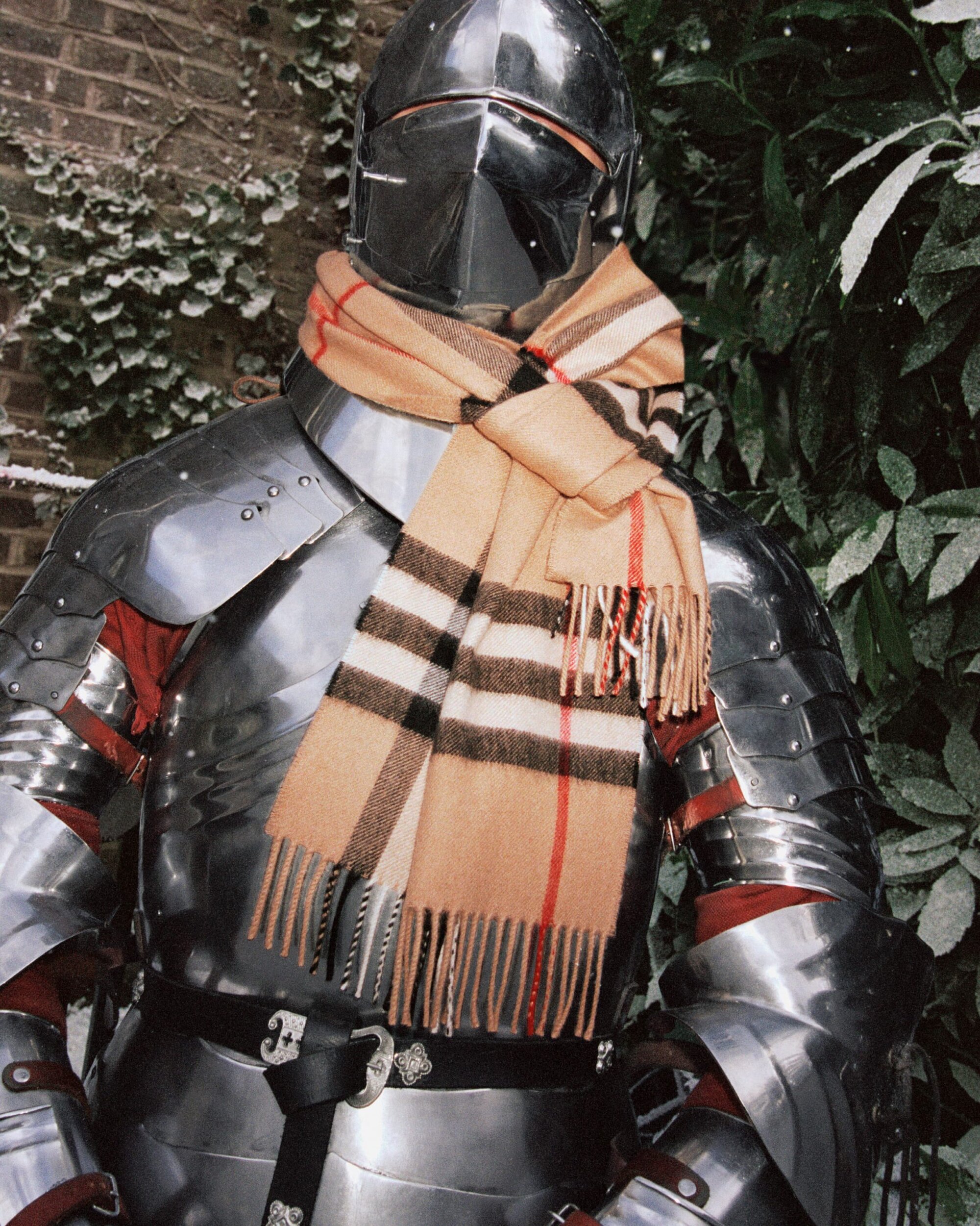 Full body shot of a person wearing shining metal armor and a tan plaid scarf in an outdoor wooded environment on a cloudy day.