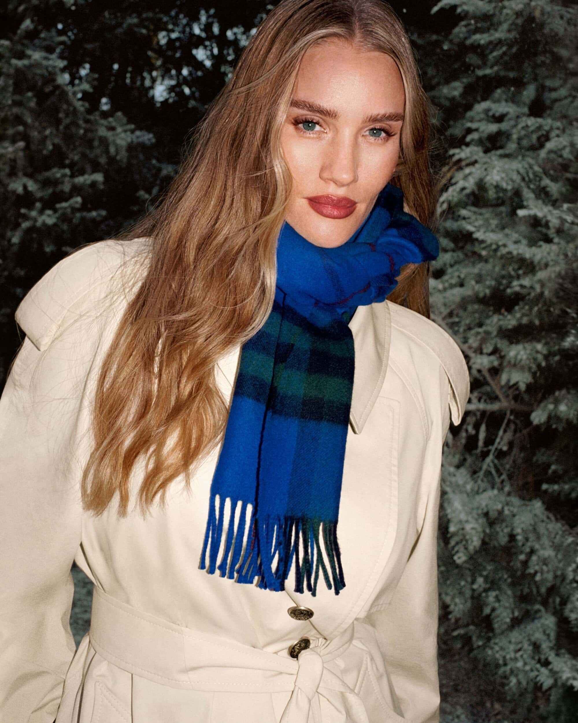 A fair-skinned adult woman poses in a light-colored coat with a blue and green scarf against an evergreen forest backdrop.