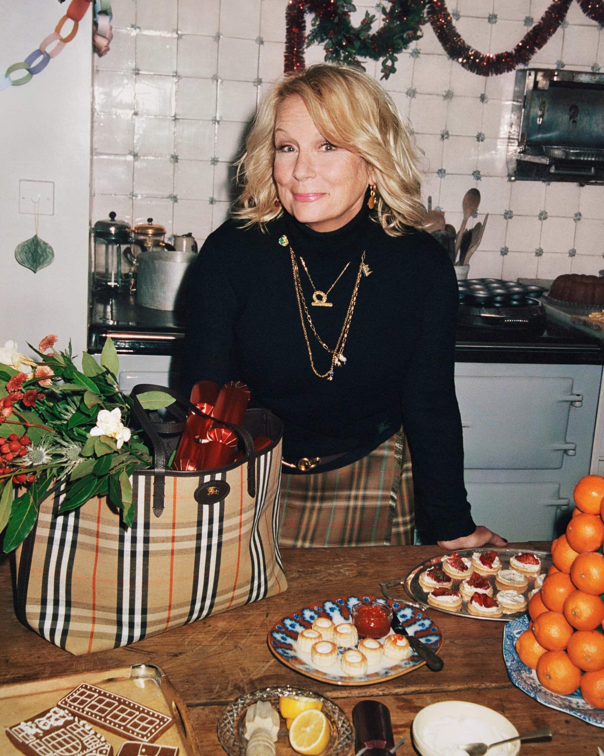 Blonde woman in a black turtleneck stands in a kitchen with food on a wooden table, including oranges, desserts, and a Burberry bag.