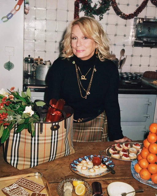Blonde woman in a black turtleneck stands in a kitchen with food on a wooden table, including oranges, desserts, and a Burberry bag.