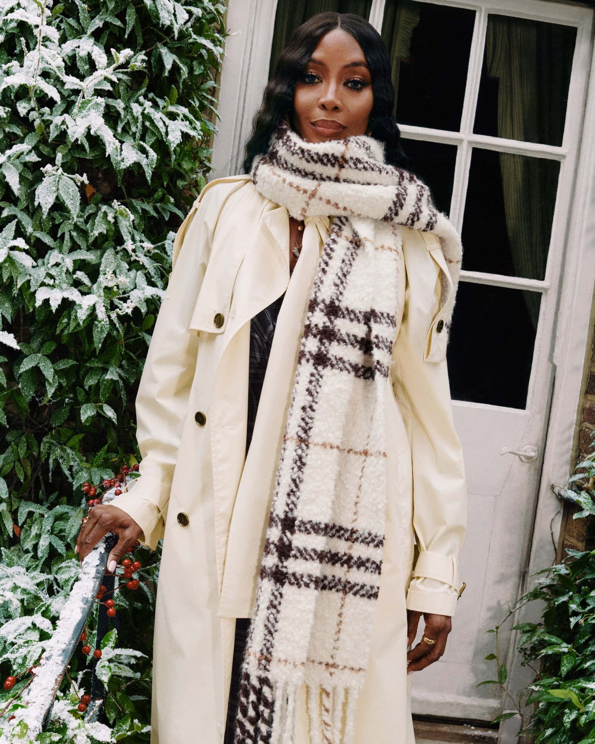 A fashionable adult female model poses outdoors in a trench coat and patterned scarf in front of a snow-covered building and greenery.