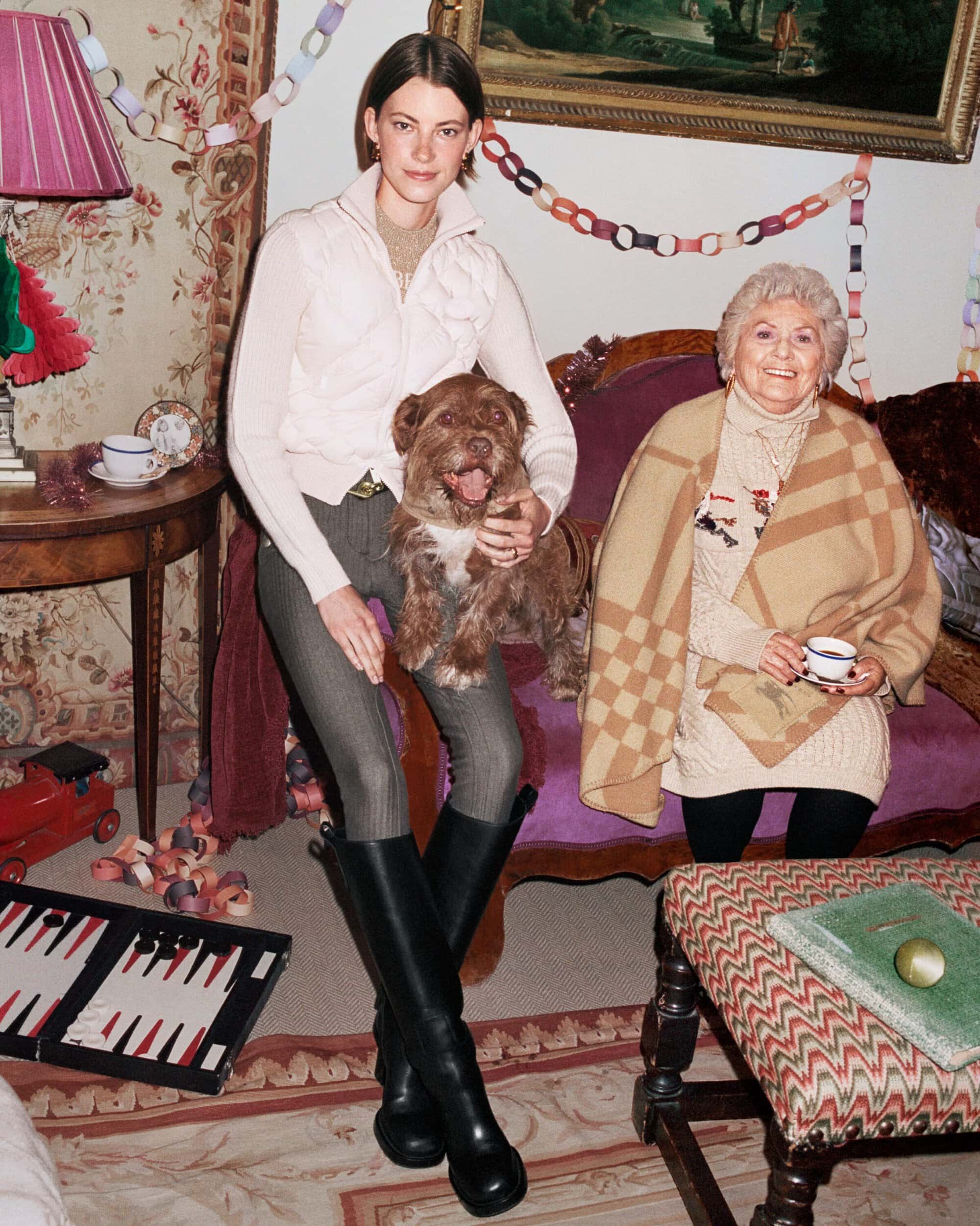 A young woman holds a dog, sitting next to an older woman on a couch, indoors. Holiday decor and backgammon board are nearby.