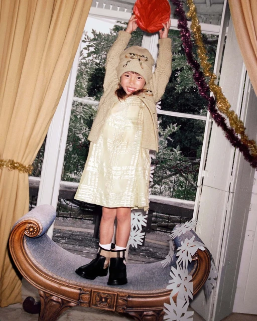 A child stands on an ornate bench indoors, holding a red ball, with natural light streaming through a window and decorations visible.