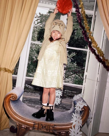 A child stands on an ornate bench indoors, holding a red ball, with natural light streaming through a window and decorations visible.