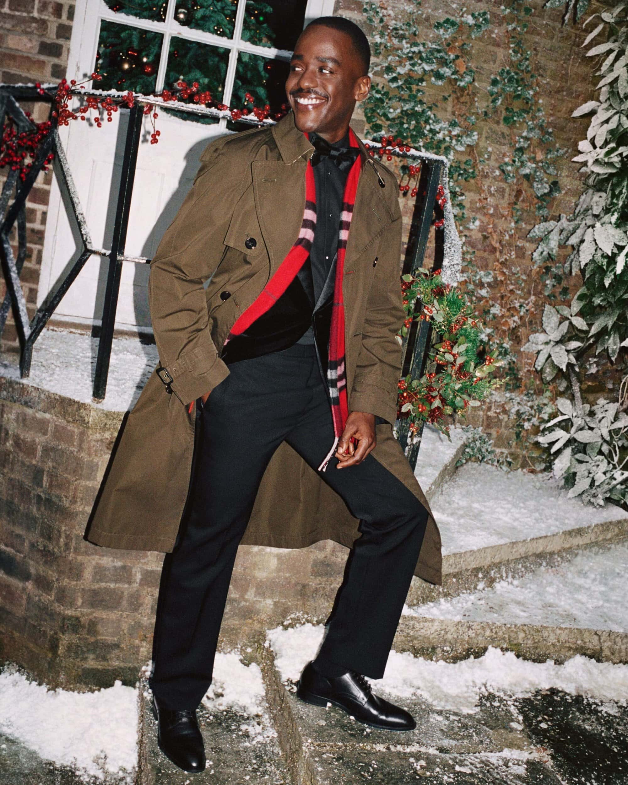 An adult man in a brown trench coat smiles and stands on snowy brick steps near a garlanded doorway outdoors during the daytime.