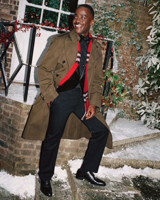 An adult man in a brown trench coat smiles and stands on snowy brick steps near a garlanded doorway outdoors during the daytime.
