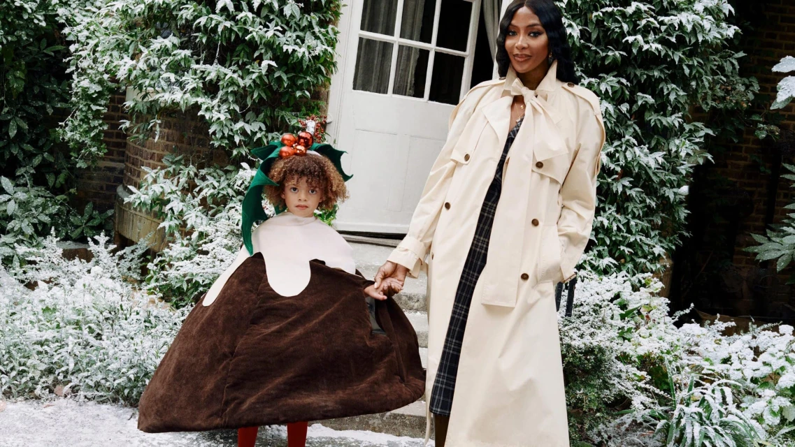 Model Naomi Campbell and a child in a Christmas pudding costume hold hands in a snow-dusted outdoor scene with greenery and a white door.