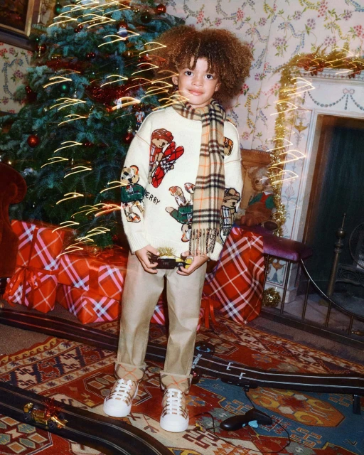 A child stands indoors in front of a Christmas tree wearing a patterned sweater, khaki pants, and white sneakers holding a toy.