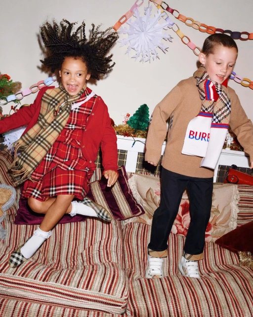 Two children, a girl with curly hair and a boy, jump on a striped couch indoors, wearing Burberry scarves and festive holiday attire.