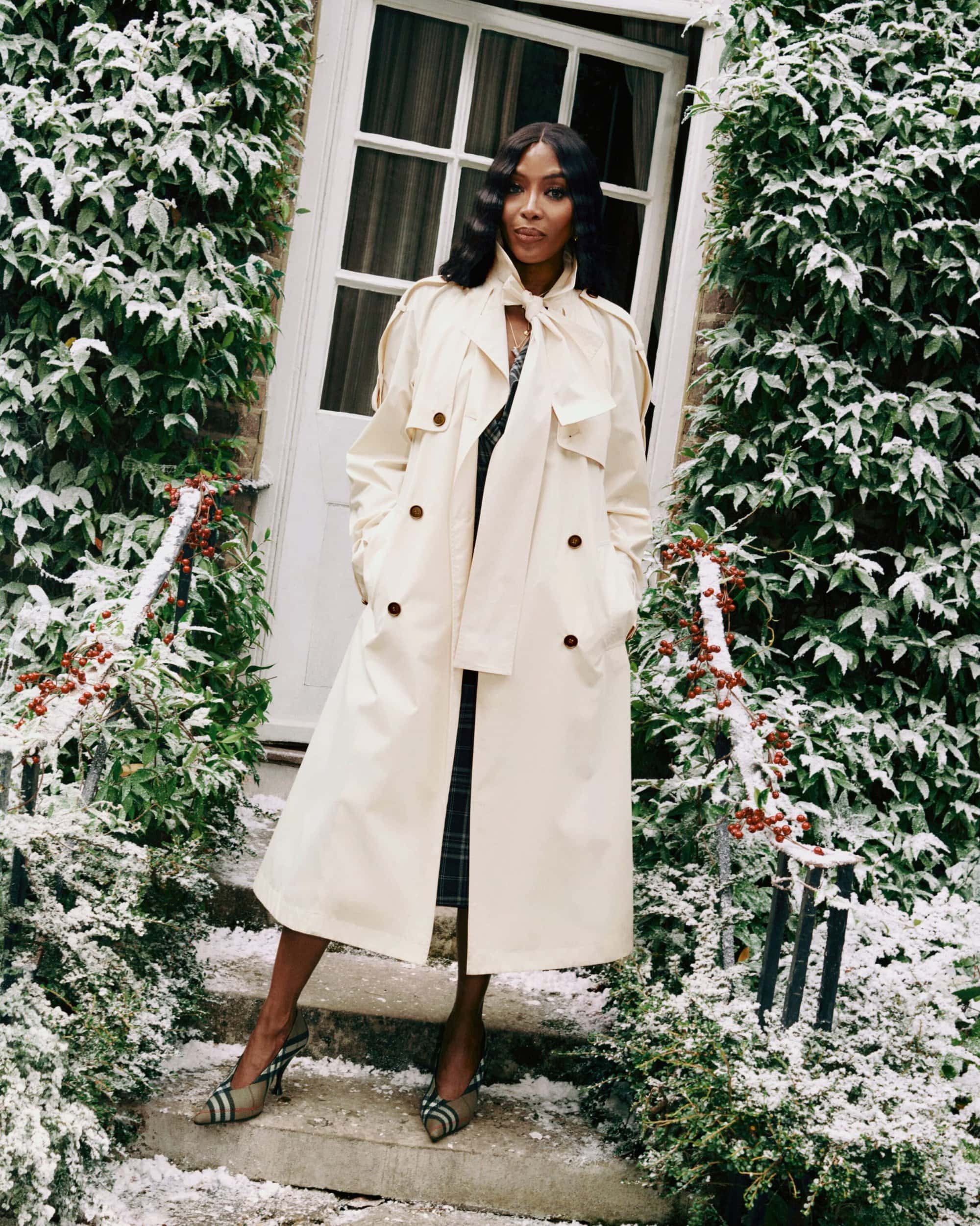 Naomi Campbell wearing a cream Burberry trench coat on snowy stone steps outside a residential building door.