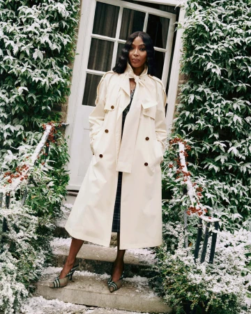 Naomi Campbell wearing a cream Burberry trench coat on snowy stone steps outside a residential building door.