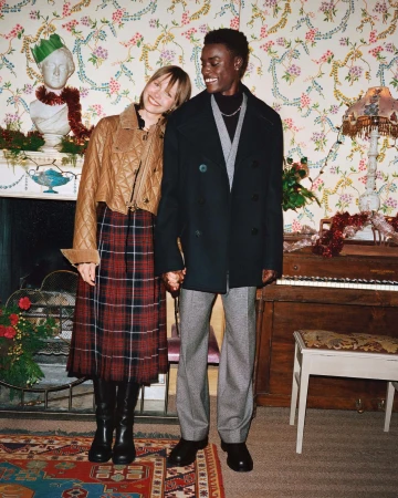 A stylish couple stands indoors holding hands, with festive decorations, a piano, and a floral wallpaper backdrop for a holiday mood.