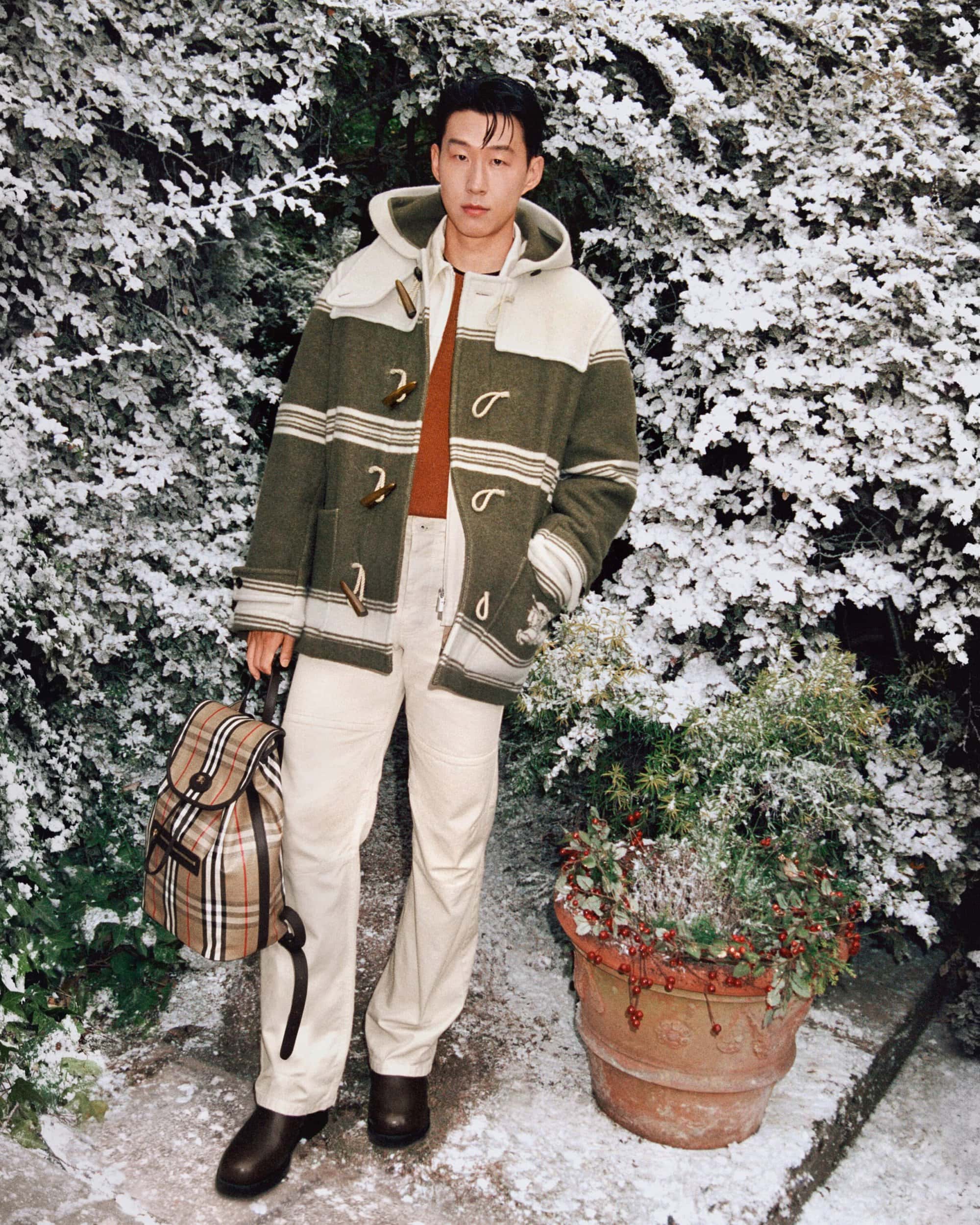 Adult male in striped duffle coat stands in a snow-dusted garden with a plaid backpack, tan pants and brown leather boots on a stone surface.