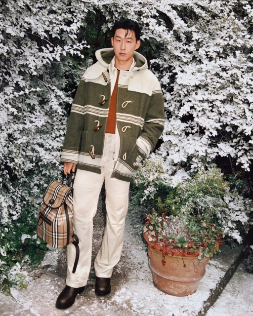 Adult male in striped duffle coat stands in a snow-dusted garden with a plaid backpack, tan pants and brown leather boots on a stone surface.
