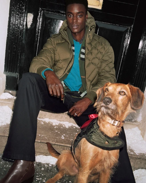 Dark-skinned man and brown dog sit on snowy steps in front of a black door wearing stylish outerwear in a medium shot.