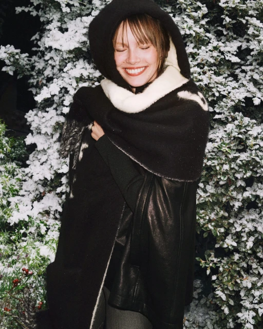 Smiling woman in black hooded poncho and winter coat standing outside in snow. Fashionable winter attire. Outdoor portrait.