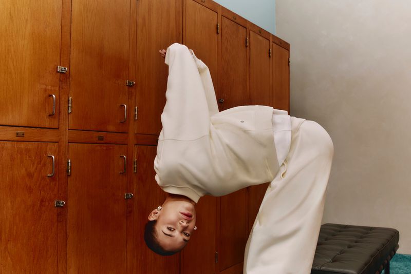 Adult woman in a white tracksuit bends over backwards in front of wooden lockers in an indoor setting with natural lighting.