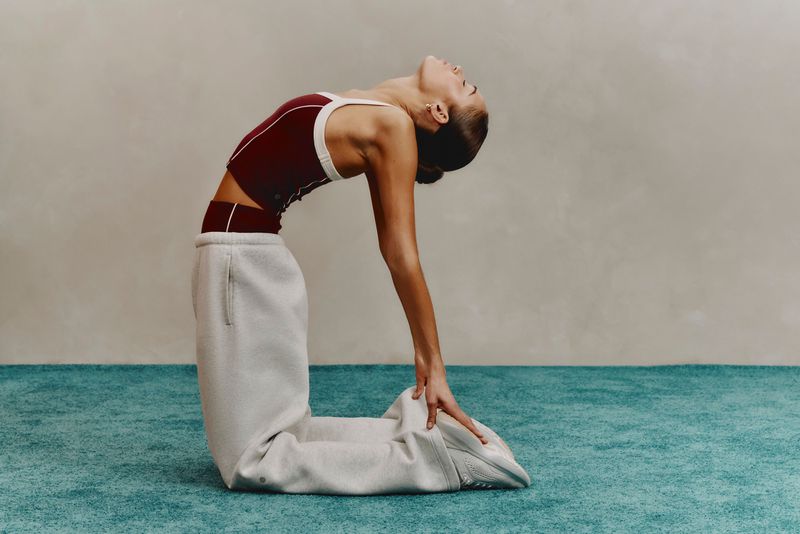 Adult female in a burgundy top and sweatpants is kneeling on a teal surface with her head tilted back in a camel pose in a studio.