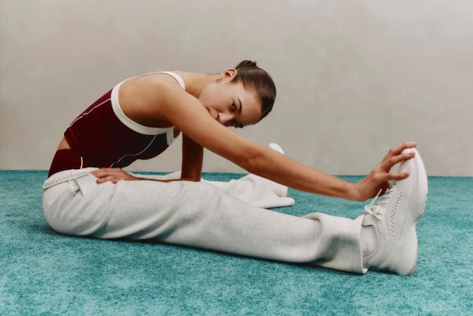 Athletic woman stretches her leg wearing a matching red and white top with white sweatpants and white sneakers in a studio setting.