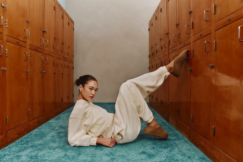 Woman in casual clothing poses on a blue carpet in between rows of wooden lockers, leaning back with her legs propped against lockers.