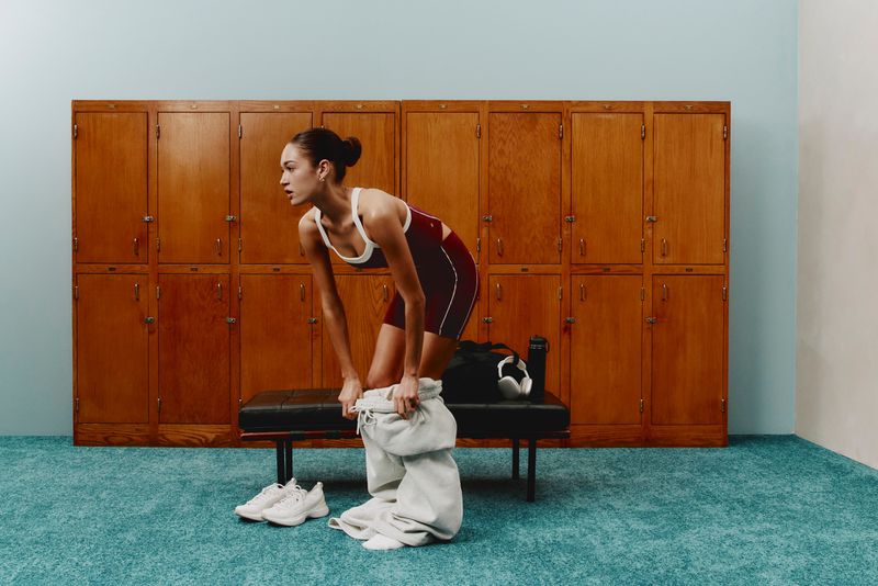 Woman in athletic wear in a locker room, adjusting pants on a bench with a gym bag and water bottle nearby, sneakers on floor.