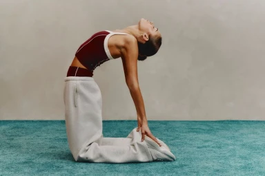 A woman in a burgundy top and grey sweatpants stretching backwards on a teal carpet against a neutral wall.