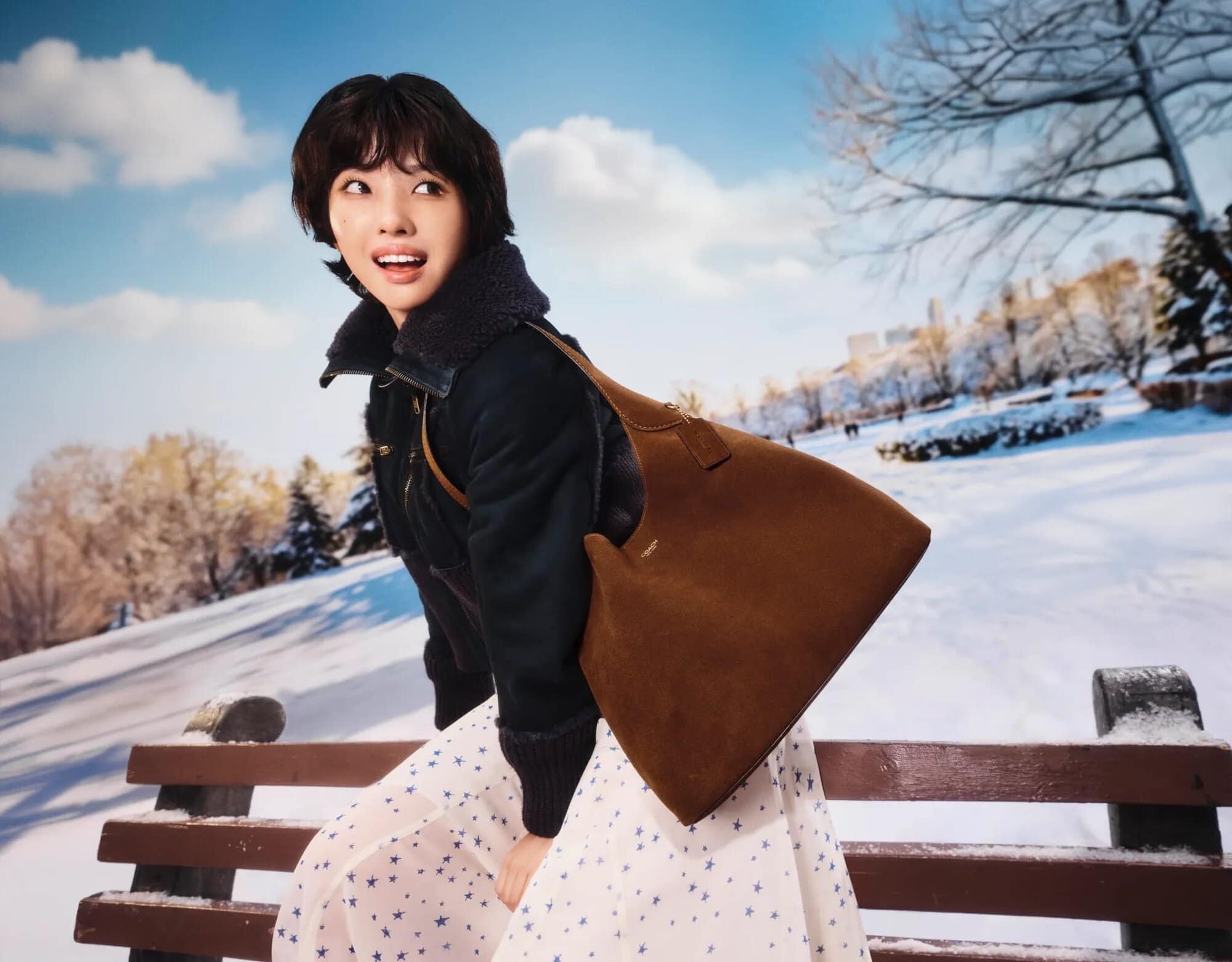 Asian woman poses with a tan leather hobo bag on a park bench in winter. The background features a snowy park landscape and bare trees.