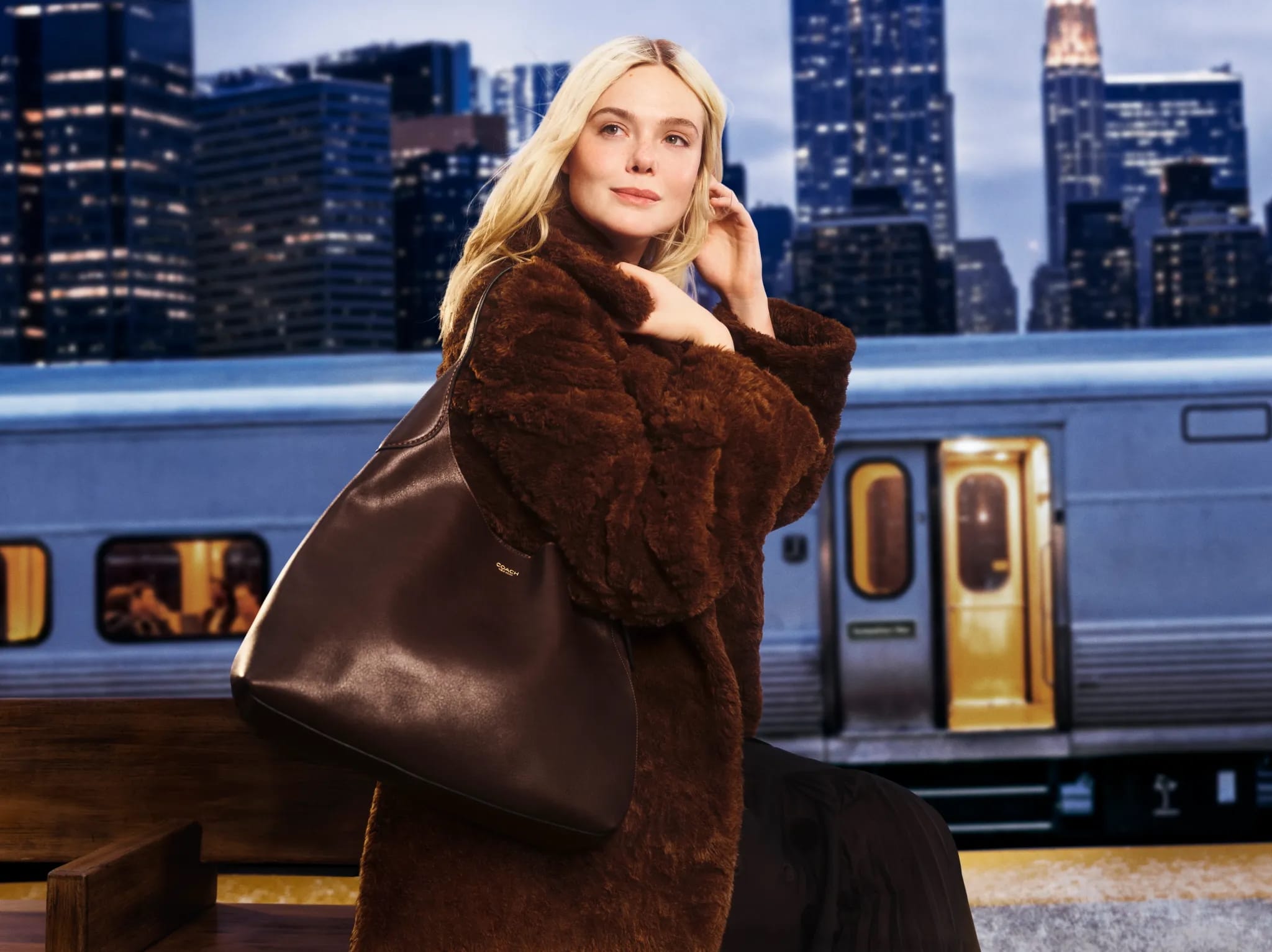 Woman in a brown fur coat posing with a brown leather bag at a train station, against a backdrop of a city skyline at night.