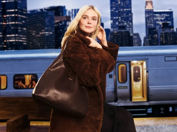 Woman in a brown fur coat posing with a brown leather bag at a train station, against a backdrop of a city skyline at night.