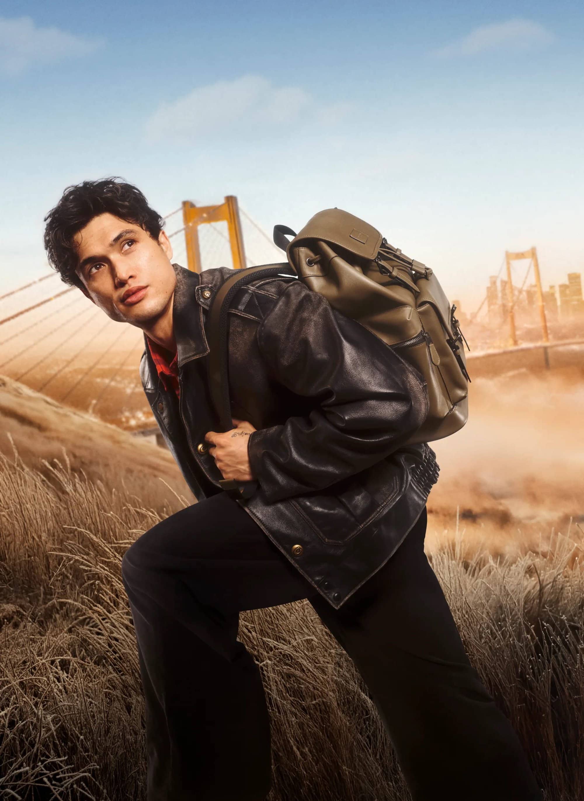 Man in a black jacket with a backpack climbs a grassy hill in an outdoor setting, with a distant bridge and city skyline.