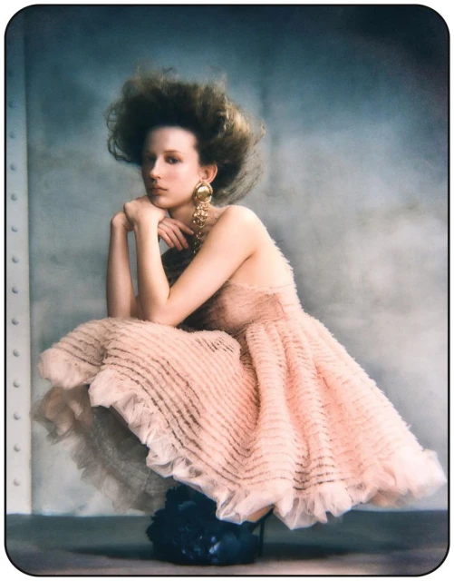 Woman in a layered pink dress posing in a crouched position against a textured gray background, showcasing fashion and portrait photography.