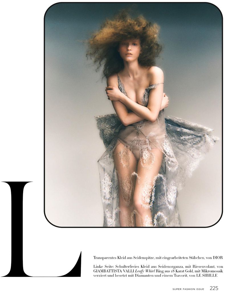 Fashion editorial shot of a slender female model with curly hair, posing in a semi-transparent lace dress with muted grey tones.